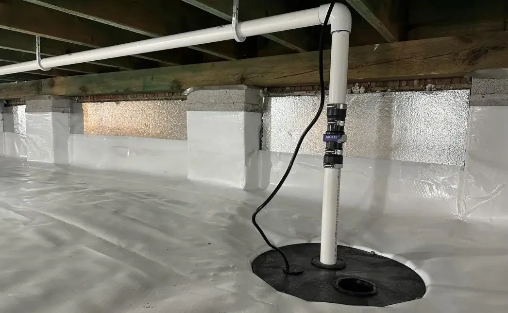 Sump pump basin with a white discharge pipe and power cord in a crawl space lined with a white vapor barrier