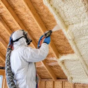 Technician spraying foam insulation onto the underside of an attic crawlspace between exposed wooden rafters.